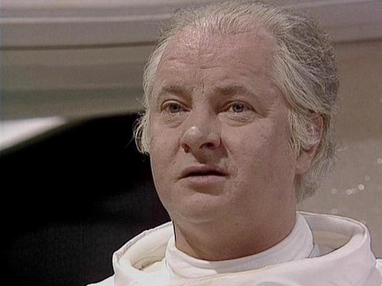 Claude 3.7 describes the image as: "The image appears to be a still from a television series, showing a person with light-colored hair wearing what looks like white robes or a white uniform with a distinctive collar. The individual has a somewhat pale appearance and appears to be in what might be a spacecraft or futuristic setting, based on the background elements.

The costume suggests this might be from a science fiction production, with the white garment possibly indicating a specific role o…