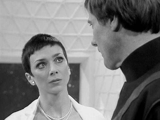 Claude 3.7 describes the image as: "This black and white image shows a scene with two people in conversation. The person on the left has a very short, pixie-style haircut and is wearing what appears to be a light-colored outfit with a pearl necklace. They have an intense, contemplative expression while looking up at the other person. The individual on the right, shown in profile, has darker hair and appears to be wearing a dark high-collared garment.

The setting features a geometric patterned …