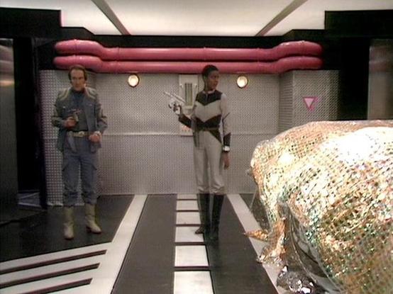 Claude 3.7 describes the image as: "This image shows a scene from a science fiction television series, set in what appears to be a futuristic spacecraft or space station interior. Two crew members are visible in a corridor or room with metallic walls and distinctive floor markings. They're wearing different uniforms - one in a grayish jumpsuit and the other in a white and dark outfit with boots. Both are holding what appear to be futuristic weapons. On the right side of the image is an object c…