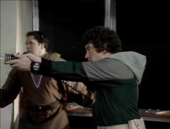 Claude 3.7 describes the image as: "The image shows a tense scene from what appears to be a science fiction television series from the late 1970s or early 1980s, based on the production quality and visual style. 

In the frame, two individuals are in what looks like a spacecraft or control room setting. One person wearing a brown outfit stands to the left, while another in a green and gray outfit is pointing what appears to be a handheld device or weapon. The scene suggests a confrontation or u…