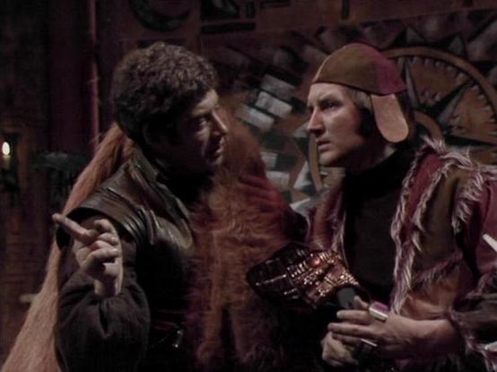 Claude 3.7 describes the image as: "This image shows a scene from a science fiction television series, featuring two characters in a dimly lit, industrial-looking setting. The scene appears to take place in some kind of mechanical or technological environment, with gears visible in the background.

The two figures are dressed in distinctive costume pieces that include fur-trimmed garments and leather-like materials. The person on the right is wearing what looks like a cap with ear flaps in a re…