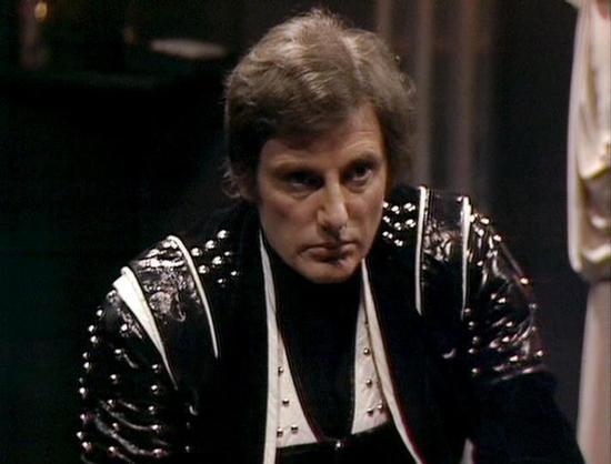 Claude 3.7 describes the image as: "This image appears to be from a science fiction television series from the late 1970s or early 1980s. It shows a person wearing a distinctive costume featuring a black leather jacket with studded shoulders and white accent panels. The costume has a somewhat militaristic or futuristic design typical of sci-fi productions from that era.

The person is wearing a dark shirt underneath the elaborate jacket and has a serious, intense expression. The setting appears…