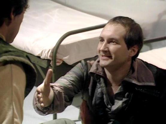 Claude 3.7 describes the image as: "This image appears to be from a science fiction television series, showing two people in what looks like a spacecraft or space station interior. The scene takes place in what appears to be living quarters with minimalist bunks or beds visible with metal frames.

One person is wearing a brown leather-type jacket and appears to be reaching out with their hand in a friendly or conversational gesture while sitting or leaning on one of the bunks. They have short d…