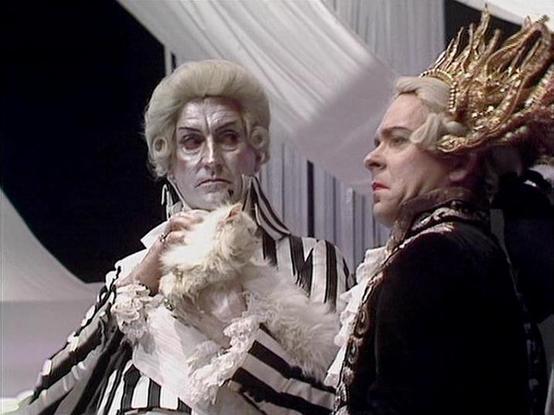 Claude 3.7 describes the image as: "This image appears to be from a period costume drama, featuring two performers in elaborate 18th-century style attire. On the left, a figure wears a distinctive black and white striped outfit with a white powdered wig and ruffled cravat. On the right, another figure is dressed in ornate dark clothing with a dramatic golden crown or headdress. The setting includes white fabric draping in the background, suggesting a formal or theatrical environment. The costum…