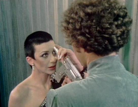 Claude 3.7 describes the image as: "The image shows a tense scene from a television series with a dramatic confrontation. One person with short dark hair and elegant makeup is wearing what appears to be formal attire with jewelry, including earrings. They are facing another person with curly hair wearing what looks like a grayish-green jacket, who is holding what appears to be a weapon or device.

The setting features light-colored curtains or fabric in the background, giving it a retro sci-fi …