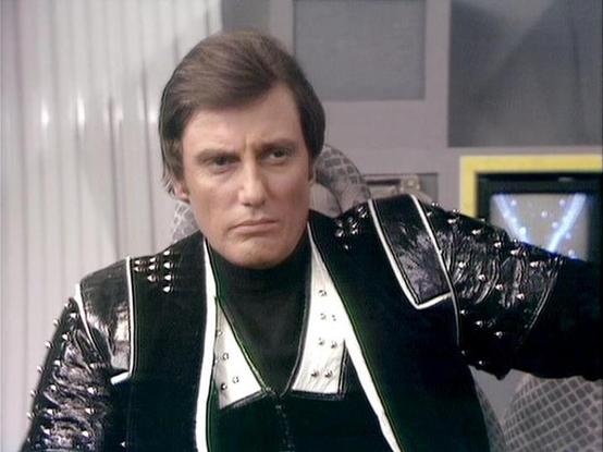 Claude 3.7 describes the image as: "The image shows a person in a distinctive costume from what appears to be a science fiction television production from the late 1970s or early 1980s. The individual is wearing a striking black leather outfit with metallic embellishments, studded details, and contrasting white panels. They have dark hair and are shown in what looks like a futuristic interior setting with light-colored walls and some technological elements visible in the background.

The costum…