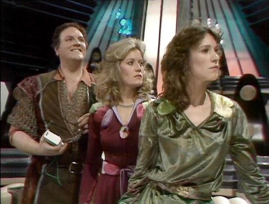 Claude 3.7 describes the image as: "The image shows a scene from a science fiction television series, appearing to be from the late 1970s or early 1980s based on the production style. Three people are standing together in what looks like a futuristic spacecraft or control room, with dramatic lighting and technical elements visible in the background.

The trio is dressed in distinctive retro sci-fi costumes - the person on the left wears a patterned vest-like garment with earthy tones, the middl…