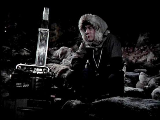 Claude 3.7 describes the image as: "This image appears to be from a dark, atmospheric scene set in what looks like a cold, possibly arctic or wilderness environment. The person in the frame is wearing a fur-lined hood parka and appears to be in a survival situation. Next to them is what looks like scientific or radio equipment with a vertical antenna or measuring device.

The setting suggests this might be depicting an expedition, research mission, or someone stranded in harsh conditions. The e…