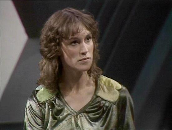 Claude Sonnet 4.0 describes the image as: "I can see this is a scene from a science fiction television series, showing a person with curly brown hair wearing a distinctive green/olive colored outfit with a metallic or satin-like finish. The costume has a futuristic design typical of 1970s-80s sci-fi productions, with a v-neck and collar styling. The setting appears to be inside a spacecraft or space station, with angular metallic walls and geometric patterns visible in the background. The light…