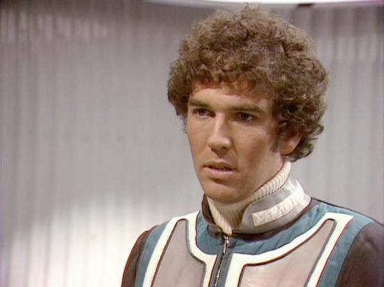 Claude 3.7 describes the image as: "The image shows a person with curly brown hair wearing a distinctive costume that features a high collar with white trim and a blue and white paneled design. This appears to be from a science fiction television production from the late 1970s or early 1980s based on the visual style and production quality. 

The costume has a futuristic appearance typical of space-based sci-fi shows of that era, with clean lines and color blocking. The background appears to be…