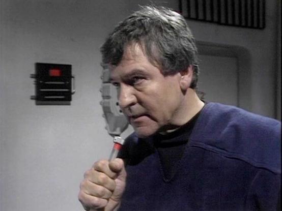 Claude Sonnet 4.0 describes the image as: "I can see this is a scene set in what appears to be a spacecraft or space station interior, with characteristic white/gray walls and technical equipment visible in the background. The person in the image is wearing a dark blue or navy colored uniform-style garment and is holding what looks like a communication device or small handheld instrument. The setting has the distinctive futuristic aesthetic typical of science fiction productions from this era, …