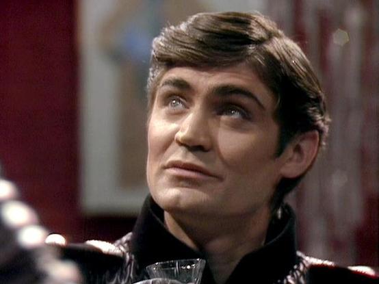 Claude Sonnet 4.0 describes the image as: "I can see this is a scene featuring a character in what appears to be a dark military or authoritarian-style uniform with distinctive collar details. The setting has a futuristic, institutional feel with blurred background elements that suggest a spaceship or command facility interior. The lighting creates a dramatic atmosphere typical of science fiction television productions from this era. The character appears to be looking upward with an intense, c…