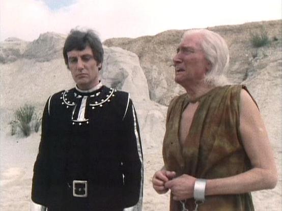 Claude 3.7 describes the image as: "The image shows a scene set in a desert or rocky landscape with light-colored formations in the background. Two figures appear to be in costume for a science fiction production. On the left is a person wearing an ornate black outfit with decorative elements around the collar and a belt with a square buckle. On the right is an older figure with light-colored hair wearing a sleeveless brown/tan tunic and what appears to be a silver bracelet or cuff.

The costum…