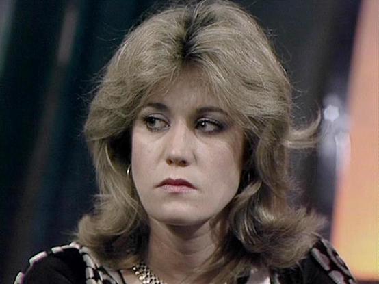 Claude 3.7 describes the image as: "The image shows a person with blonde, feathered hair styled in a popular 1970s/early 1980s fashion. They're wearing what appears to be a patterned top with a necklace, and have a serious or contemplative expression. The lighting and film quality suggest this is from a television production of that era, with a somewhat dark background and dramatic lighting typical of science fiction or drama productions from that time period. The styling, costume and productio…