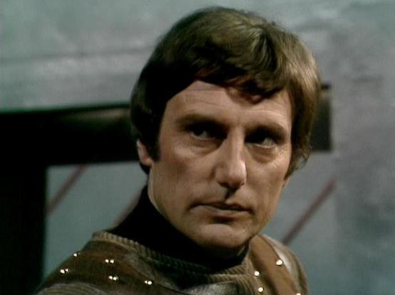 Claude 3.7 describes the image as: "The image shows a person in what appears to be a science fiction setting, with a muted, industrial-looking background. They're wearing what looks like a brown costume with some reflective or shiny elements, possibly part of a uniform or specialized outfit typical of vintage sci-fi productions. The lighting and film quality suggest this is from a television production from the 1970s or early 1980s era, which is characteristic of classic British science fiction…