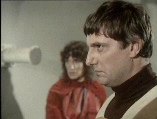 Claude Sonnet 4.0 describes the image as: "I can see this appears to be a scene from a science fiction television series, showing two people in what looks like a futuristic or spaceship-like setting with white/light colored walls in the background. One person is wearing what appears to be a red or burgundy colored outfit, while the other is wearing darker clothing with what looks like a light-colored vest or jacket over it. The lighting and film quality suggests this is from a television produc…
