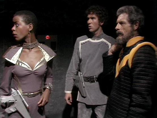 Claude Sonnet 4.0 describes the image as: "I can see this is a scene featuring three characters in what appears to be a futuristic or sci-fi setting with dark atmospheric lighting. The characters are wearing distinctive costumes - one person is dressed in form-fitting attire with metallic armor-like details and decorative elements, another is wearing a gray tunic-style outfit, and the third is in dark clothing with yellow/orange trim or sash details. The lighting and staging suggests this is fr…