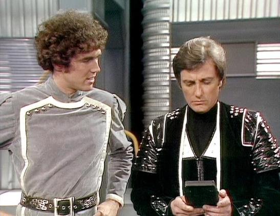 Claude Sonnet 4.0 describes the image as: "This scene appears to be set aboard a spaceship, likely the Liberator, with its characteristic futuristic interior featuring metallic surfaces and technological elements. Two characters are shown in conversation - one wearing a light grey military-style uniform with decorative elements and a belt, while the other is dressed in a distinctive black leather outfit with metallic studs and trim, holding what appears to be a small device or communicator. The…