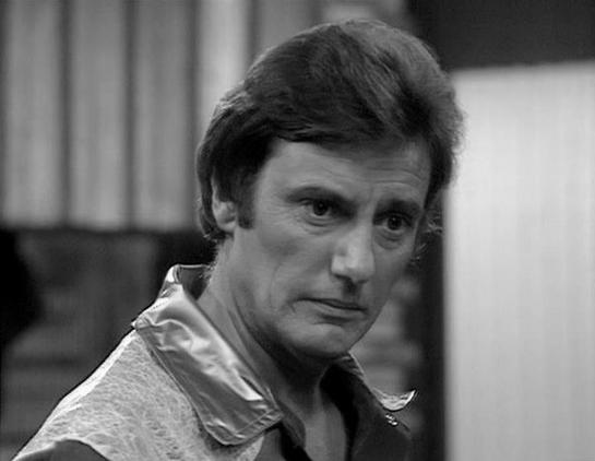 Claude Sonnet 4.0 describes the image as: "I can see this is a black and white photograph showing a man with dark, styled hair wearing what appears to be a light-colored shirt or jacket. The image has a dramatic, moody quality typical of science fiction television from the late 1970s or early 1980s. The lighting and composition create shadows that emphasize the subject's facial features. The background appears to show some kind of interior setting, though it's blurred and out of focus. The over…