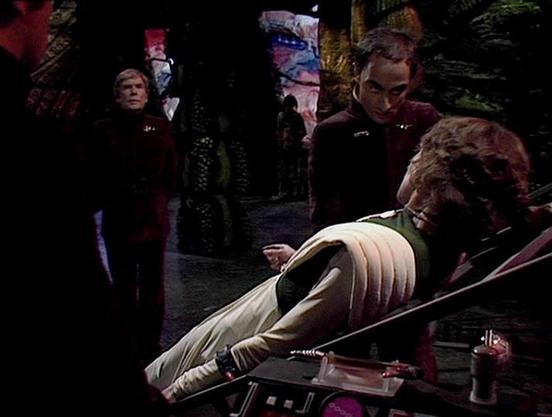 Claude 3.7 describes the image as: "This image appears to be from a science fiction television series, showing a scene in what looks like a futuristic or alien setting. There are several people in the scene wearing distinctive uniforms or costumes with maroon/burgundy coloring. In the foreground, someone is lying on what appears to be a medical bed or examination table, wearing a ribbed or padded outfit with light coloring. Another person in a maroon uniform seems to be attending to them. The b…