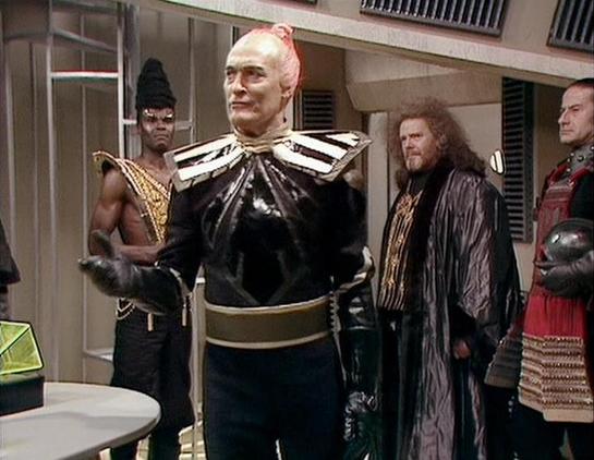 Claude 3.7 describes the image as: "This image shows a scene from a science fiction television production featuring several characters in distinctive costumes. In the center is a figure with a striking appearance, wearing a black outfit with pronounced shoulders and a gold belt. They have pale makeup and reddish hair styled upward. Surrounding this central character are others in varied futuristic attire - one in minimal black clothing with gold accents, another in dark robes with curly hair, a…