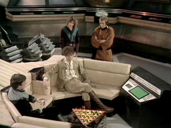 Claude 3.7 describes the image as: "This image shows a scene set in what appears to be a spaceship interior with a futuristic design. Four individuals are pictured in a lounge-like area with cream-colored seating arranged in a sunken conversation pit style. There's a colorful triangular game board on a table in the foreground.

The setting features typical sci-fi elements of the late 1970s/early 1980s era television production, with distinctive costume styling of that period - including brown a…