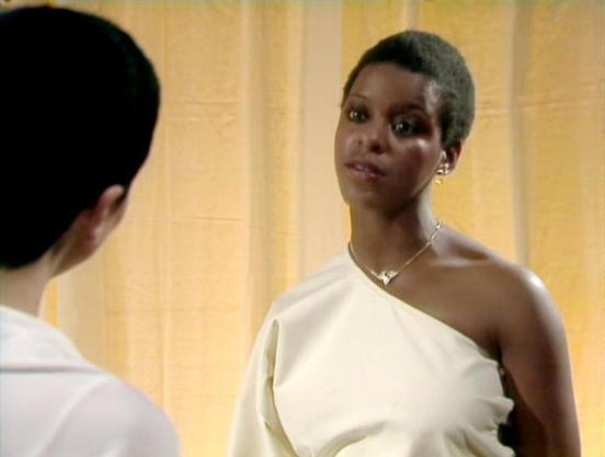 Claude 3.7 describes the image as: "The image shows a scene with two people in conversation. The person on the right is wearing an elegant one-shoulder cream or white outfit with a delicate necklace and has a short hairstyle. They appear to be in a room with warm yellow or beige walls in the background.

The scene seems to be from a dramatic moment, possibly a formal or important encounter between the characters. The lighting and setting suggest this might be from a science fiction or drama ser…