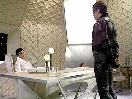 Claude Sonnet 4.0 describes the image as: "I can see this appears to be from a science fiction television series, showing a futuristic interior setting with distinctive geometric wall patterns and modern furniture. The scene takes place in what looks like a sleek, sterile environment with white and metallic surfaces. There's a desk or table in the foreground, and the setting has the characteristic design aesthetic of 1970s/80s sci-fi productions - clean lines, angular patterns, and a minimalist…