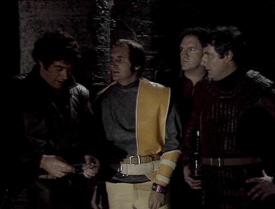 Claude 3.7 describes the image as: "This image appears to be from a science fiction television series, showing a dark, moody scene with four individuals standing together in what looks like a spacecraft or futuristic setting. The scene has a distinctly retro sci-fi aesthetic typical of 1970s-80s British television productions.

The characters are wearing distinctive costumes - one in a yellow/beige outfit with some type of arm support or design element, while others are dressed in darker leathe…