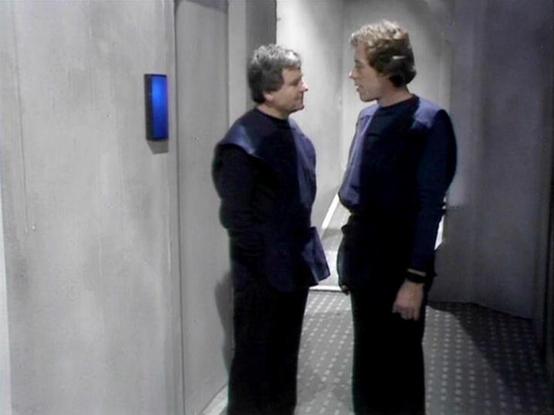 Claude 3.7 describes the image as: "This image shows two individuals having a conversation in a stark, futuristic corridor. They're both wearing similar dark uniforms with blue shoulder sections, typical of the science fiction aesthetic of the late 1970s/early 1980s British television production. The minimalist set features gray walls with a small blue panel visible in the background, and a textured floor. The corridor has the institutional, utilitarian look common in sci-fi productions of that…