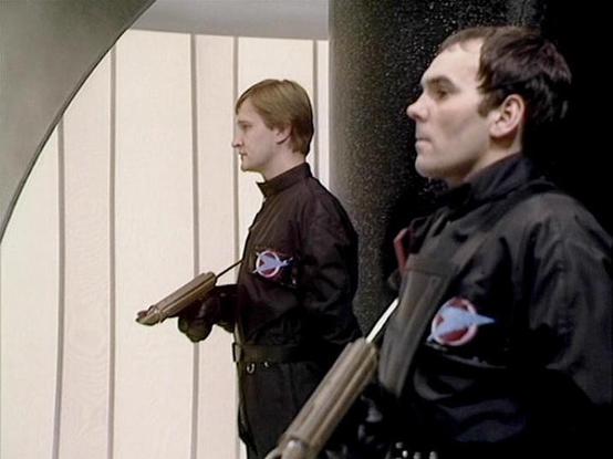 Claude Sonnet 4.0 describes the image as: "I can see two men in dark military or security-style uniforms with patches or insignia on their sleeves. They appear to be holding weapons and are positioned in what looks like a futuristic corridor or interior space with clean, geometric architecture featuring white walls and angular design elements. The setting has a sterile, institutional appearance typical of science fiction productions. The men appear to be in some kind of guard or patrol role bas…