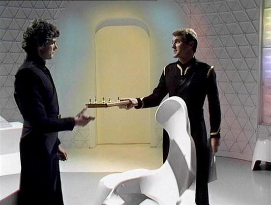 Claude 3.7 describes the image as: "The image shows a scene from a science fiction television series set in a futuristic, minimalist environment with a distinctive honeycomb-patterned wall. Two individuals in dark uniforms appear to be exchanging what looks like a chess set or similar game board. The setting has a clean, sterile aesthetic typical of 1970s-80s British sci-fi productions, with a white sculptural chair visible in the foreground. The room features soft lighting and a doorway with a…
