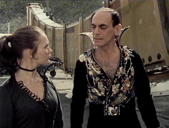 Claude 3.7 describes the image as: "This image appears to be from a science fiction television series, showing two people in conversation. On the left is a person with dark hair pulled back, wearing a black outfit with a choker necklace. On the right is a person in a distinctive costume featuring a black shirt with a floral pattern vest or overlay and an unusual collar design.

The scene looks to be taking place in what appears to be an industrial or spacecraft setting, with metal structures vi…