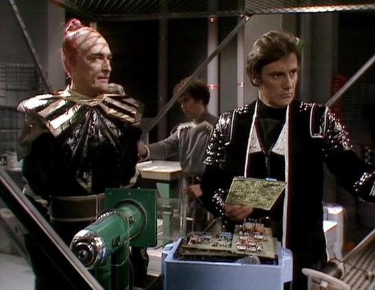Claude 3.7 describes the image as: "This image shows a scene from a science fiction television production from the late 1970s or early 1980s, based on the distinctive costume design and set aesthetics. The scene takes place in what appears to be a futuristic control room or spacecraft interior.

Two figures are prominently featured wearing elaborate costumes - one on the left in a metallic, gold-toned outfit with a distinctive headpiece, and one on the right in a black uniform with shiny detail…
