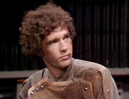 Claude 3.7 describes the image as: "This image appears to be from a science fiction television production from the late 1970s or early 1980s based on the visual style and costume design. 

The scene features a person with distinctive curly hair wearing what appears to be a beige or tan costume with decorative elements or fasteners visible on the shoulder area. The costume has a somewhat retro-futuristic design typical of science fiction productions from that era.

The background is dark with wh…