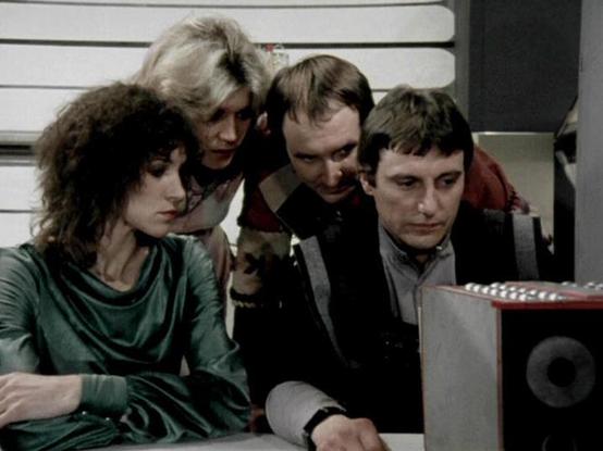 Claude Sonnet 4.0 describes the image as: "This appears to be a scene set aboard the Liberator spacecraft, showing four crew members gathered around what looks like a control console or computer terminal. The setting has the characteristic white and metallic interior design of the ship's flight deck or computer room. The group appears to be engaged in examining something on the screen or discussing mission-critical information, which was typical of scenes where the crew would analyze data, plan…