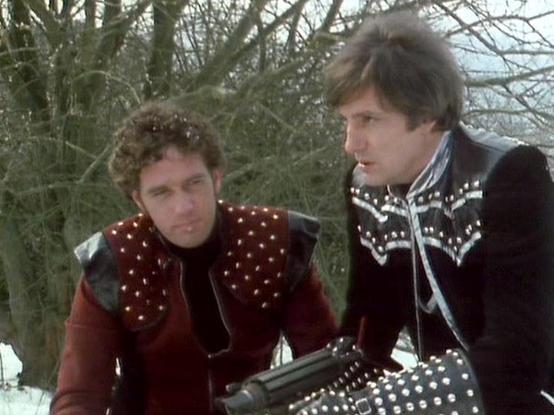 Claude 3.7 describes the image as: "This image appears to be from a retro science fiction television show, showing two people in distinctive costume design typical of late 1970s/early 1980s sci-fi aesthetic. They're wearing elaborate outfits featuring decorative studs and patterns - one in a predominantly red outfit with silver accents, and the other in black with white patterned detailing. They appear to be in a winter setting with bare trees in the background and what looks like snow on the g…
