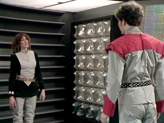Claude 3.7 describes the image as: "This image appears to be from a science fiction television series from the late 1970s or early 1980s based on the production design and costumes. The scene takes place in what looks like a futuristic interior with dark walls, metallic panels, and distinctive textured wall elements.

Two people are shown in conversation. On the left is a person with curly hair wearing a black and white outfit with a belt. On the right is someone in a distinctive 80s-style cost…