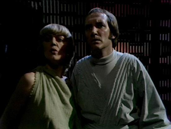 Claude Sonnet 4.0 describes the image as: "I can see two people in what appears to be a library or archive setting, with shelves of books or documents visible in the background. The scene has the distinctive lighting and production values typical of late 1970s British science fiction television. One person is wearing a light-colored, loose-fitting garment, while the other is dressed in what appears to be a turtleneck or similar top. The setting suggests this might be taking place in some kind o…