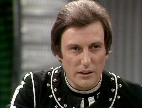 Claude Sonnet 4.0 describes the image as: "This image shows a character in a distinctive black leather outfit with metallic studs and decorative elements, typical of the futuristic costume design from late 1970s science fiction television. The character is wearing what appears to be a form-fitting black uniform with silver or chrome studs arranged in patterns across the chest and shoulders. The setting appears to be indoors with a blurred background. The costume design reflects the show's space…