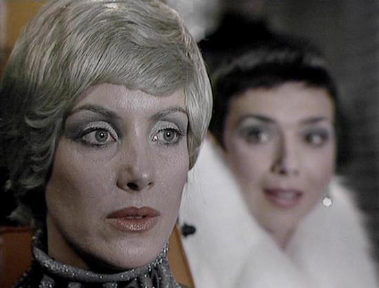 Claude Sonnet 4.0 describes the image as: "This image shows a scene with two characters in what appears to be an interior setting. The woman in the foreground has distinctive short platinum blonde hair and pale makeup, wearing what looks like a metallic or silver high-necked garment with ornate detailing. Her expression appears serious or contemplative. Behind her, slightly out of focus, is another woman with dark hair wearing what appears to be a white or light-colored outfit. The lighting and…