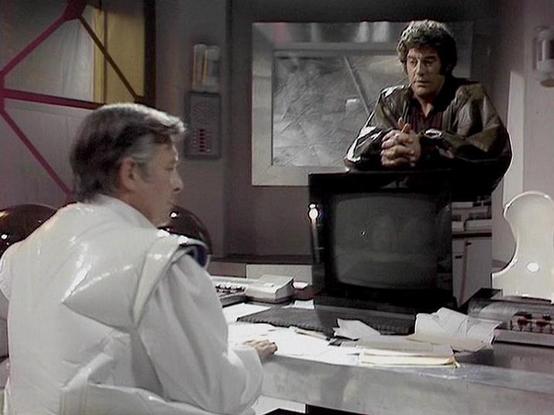 Claude 3.7 describes the image as: "This image appears to be from a science fiction television series from the late 1970s or early 1980s. The scene shows an office or control room setting with vintage technology. 

On the left, there's a person in a white uniform or coat seated at a desk with papers and equipment. On the right, there's another person in a brown leather jacket who appears on a monitor or television screen, suggesting this might be a communication between two locations.

The sett…