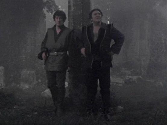 Claude 3.7 describes the image as: "This image appears to be from a vintage science fiction television series, captured in a dark, moody setting. The scene shows two figures standing together in what looks like a misty or foggy outdoor environment with ruins or deteriorated structures in the background. 

One person is wearing what appears to be a utility-style outfit with a belt, while the other is dressed in darker clothing that resembles a suit or jacket. The grainy, low-contrast quality and…