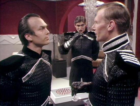 Claude Sonnet 4.0 describes the image as: "This scene takes place in what appears to be a luxurious interior setting with red and white decor. Three men are wearing dark military-style uniforms with metallic detailing and ornate trim. Two men are facing each other in the foreground in what seems to be a tense confrontation or serious discussion, while a third figure stands in the background. The costumes and set design have the distinctive futuristic aesthetic typical of late 1970s science fict…
