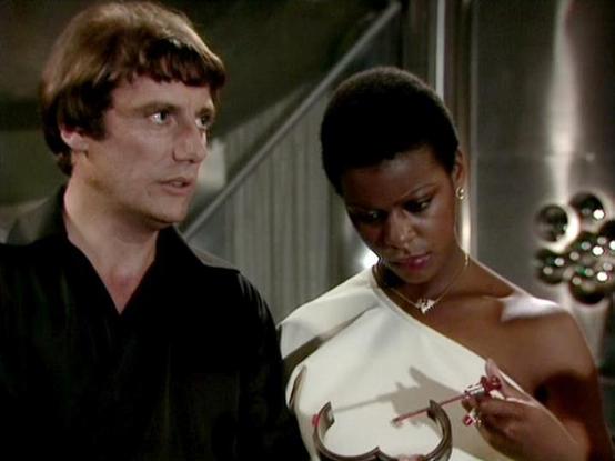 Claude 3.7 describes the image as: "This image appears to be from a science fiction TV series from the late 1970s or early 1980s based on the visual style and production quality. The scene shows two people in what looks like a spacecraft or futuristic setting, with metallic elements visible in the background. 

One person wears a black outfit while the other wears an elegant white off-shoulder garment and appears to be holding or examining something. The person on the right is adorned with a ne…