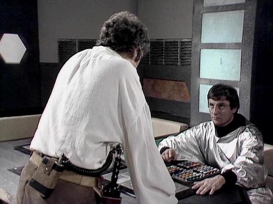 Claude Sonnet 4.0 describes the image as: "I can see two people in what appears to be a futuristic medical or technical facility setting. One person is standing, wearing white robes or garments, while another is lying down on what looks like a medical bed or examination table, wearing light-colored clothing. The background shows typical sci-fi television production design with control panels, screens, and technological equipment typical of the era. The setting has a clinical, sterile appearance…