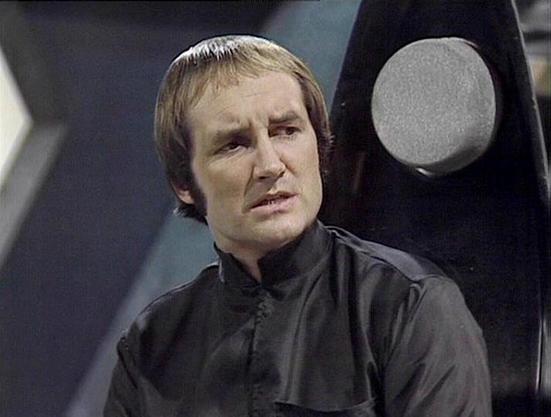 Claude 3.7 describes the image as: "This image shows a person with short hair wearing a dark, high-collared uniform in what appears to be a futuristic sci-fi setting. The person has a somewhat concerned or serious expression, and is positioned against a gray/metallic background that suggests the interior of a spacecraft or space station. 

The costume design is characteristic of 1970s-80s British science fiction television, with its minimalist styling and dark colors. The setting has the distin…