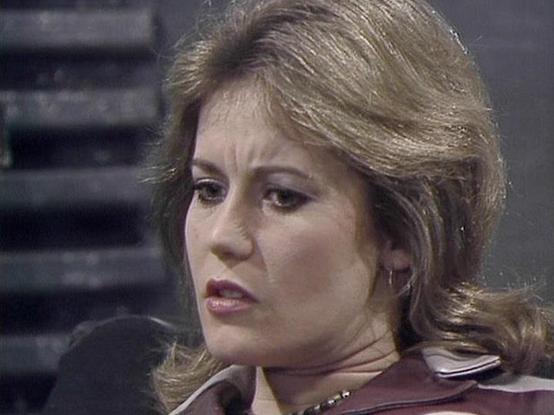 Claude 3.7 describes the image as: "The image shows a person with feathered blonde-brown hair in what appears to be a scene from a television production, likely from the late 1970s or early 1980s based on the styling and image quality. They are wearing what looks like a burgundy or reddish-brown jacket or top, and the shot is framed as a close-up showing their profile against a dark background.

The serious expression and dramatic lighting suggest this is from a science fiction or drama series.…