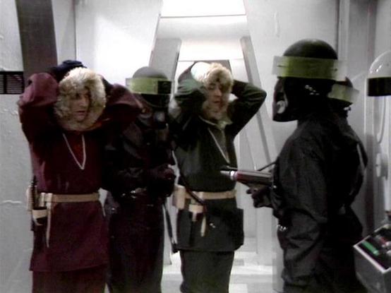 Claude 3.7 describes the image as: "This image appears to be from a science fiction television series, showing a scene in what looks like a spacecraft or space station corridor with white walls and futuristic design elements. 

The image shows several people in costume - some wearing colorful outfits in burgundy and green with distinctive headwear, while a figure on the right is dressed in dark clothing and appears to be holding what looks like a weapon. The individuals with their hands near th…