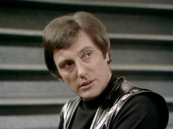 Claude 3.7 describes the image as: "This image shows a character in what appears to be a science fiction setting, wearing a distinctive black and silver costume with a high collar. The person has short brown hair and is looking slightly to the side against a background with horizontal lines or panels. The costume has a sleek, futuristic appearance typical of classic sci-fi television from the late 1970s or early 1980s. The styling, set design, and costume suggest this is from a British science …